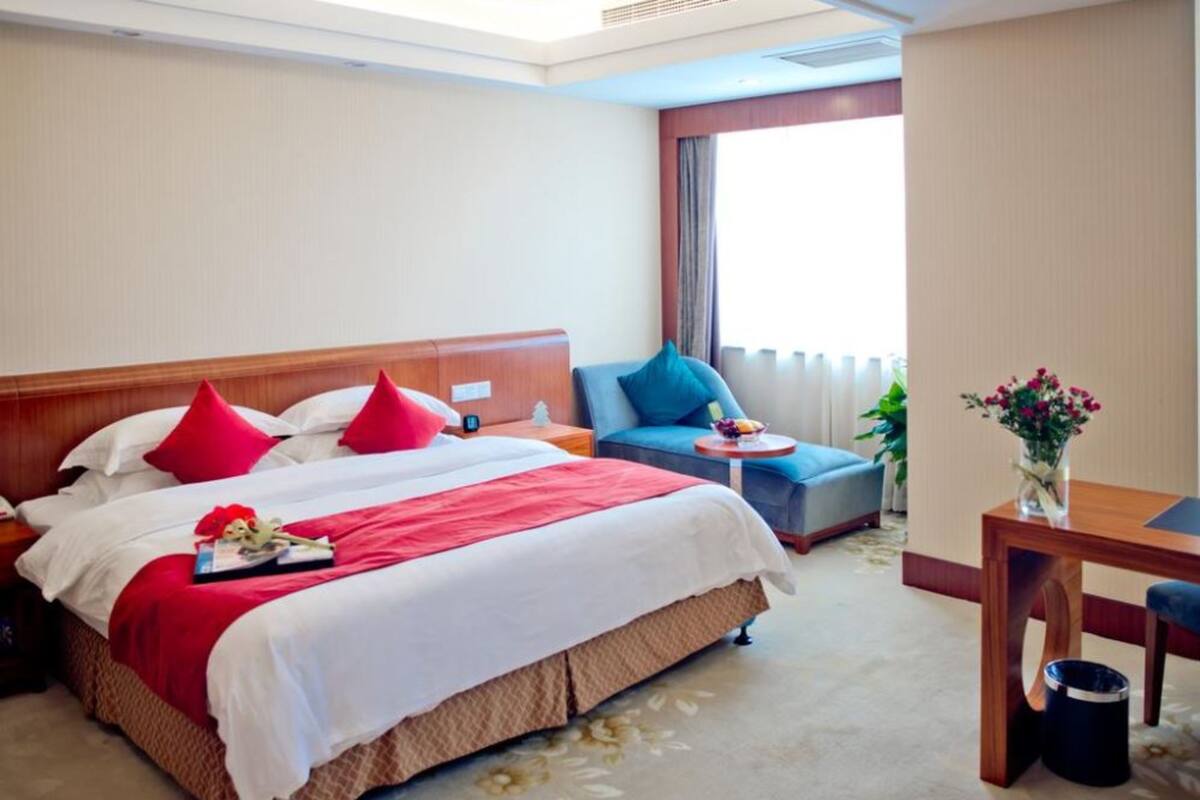 A hotel room with a large bed, a small table, a chair, and a window with curtains.