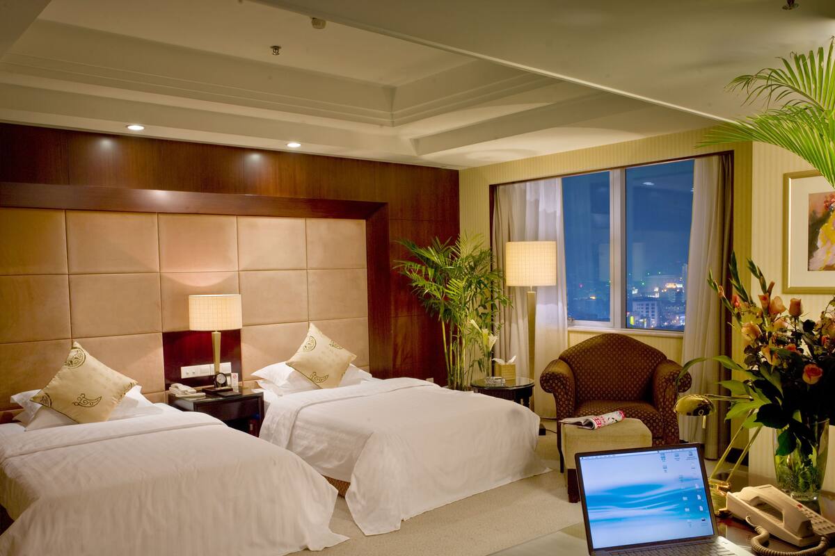 A hotel room with two beds, a desk with a laptop, a telephone, and a window with a city view.