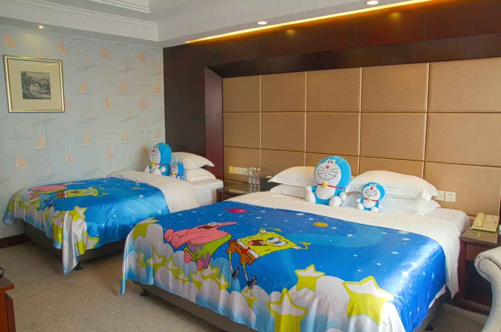 Quarto de hotel com duas camas, cada uma com colcha de temática de desenho animado, uma cabeceira grande e acolchoada, e um quadro na parede.