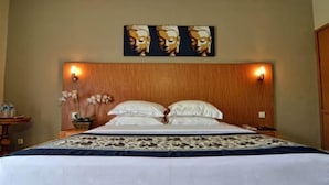 A neatly made bed with white linens and a patterned bedspread, flanked by two bedside tables with lamps. A wall-mounted artwork above the bed depicts three figures. - Taman Rosani Hotel (Keroboken Kelod)