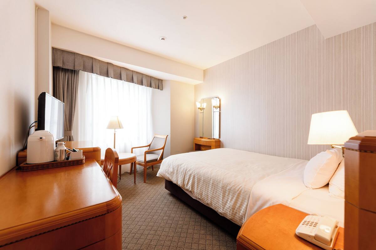 A hotel room with a bed, a desk with a lamp, a chair, a bedside table with a phone, and a window with curtains.