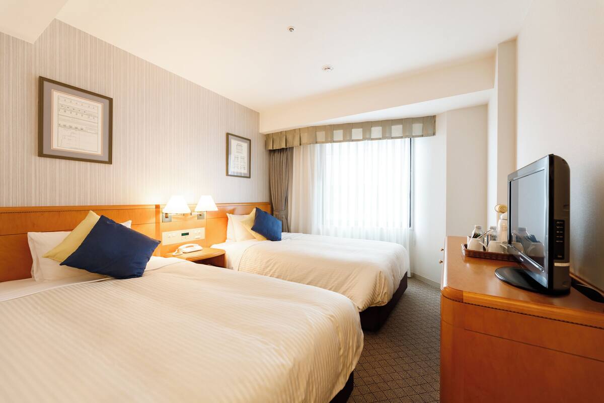 A hotel room with two beds, a television on a wooden dresser, and a window with curtains.