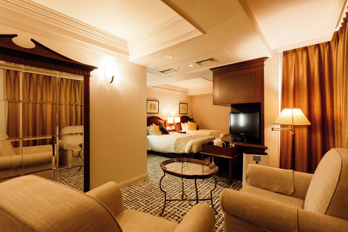 A hotel room with a bed, a TV, a sofa, a chair, a small table, and a lamp.