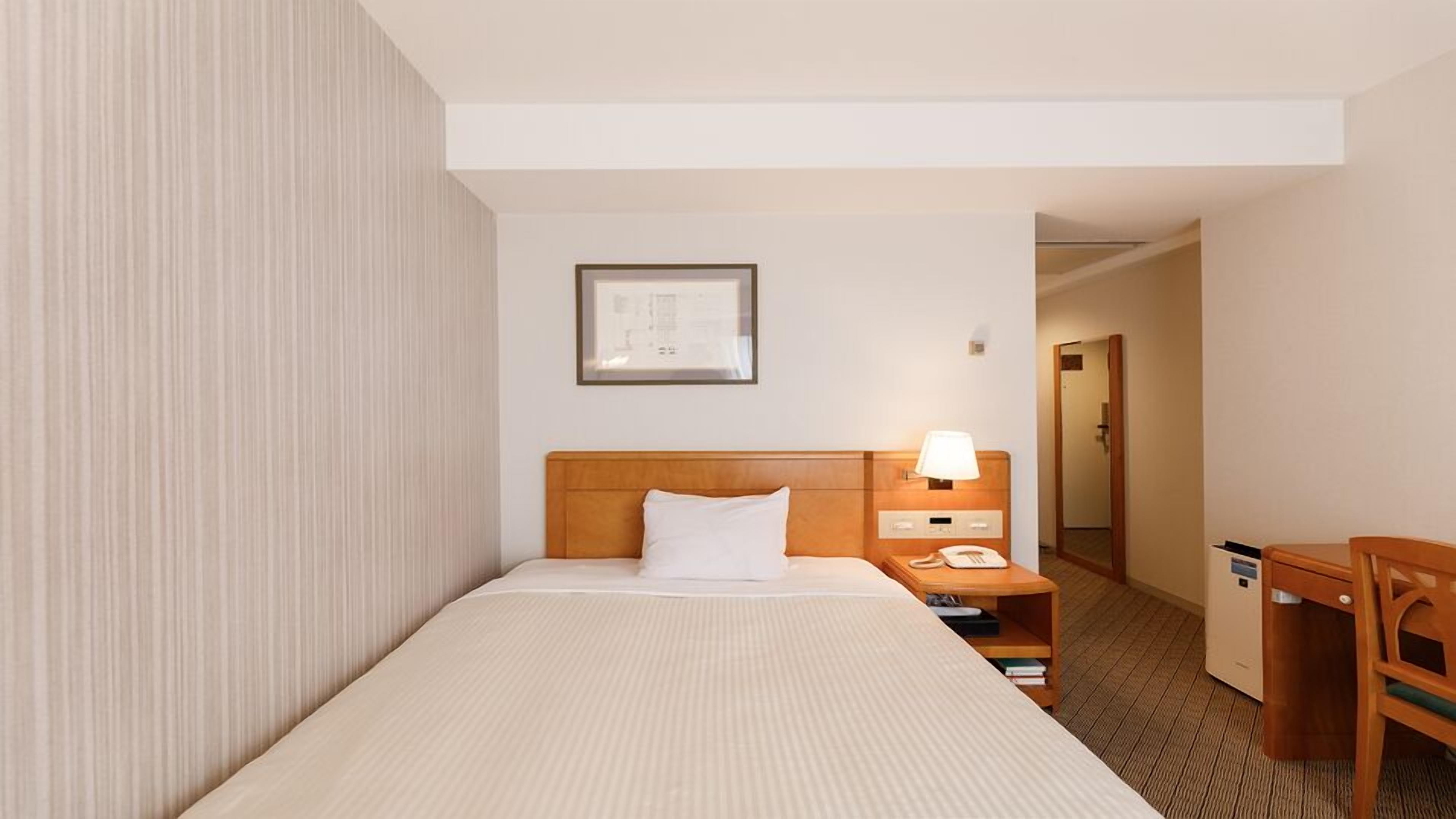 A hotel room with a single bed, a bedside table with a lamp, a phone, and a small shelf with books.