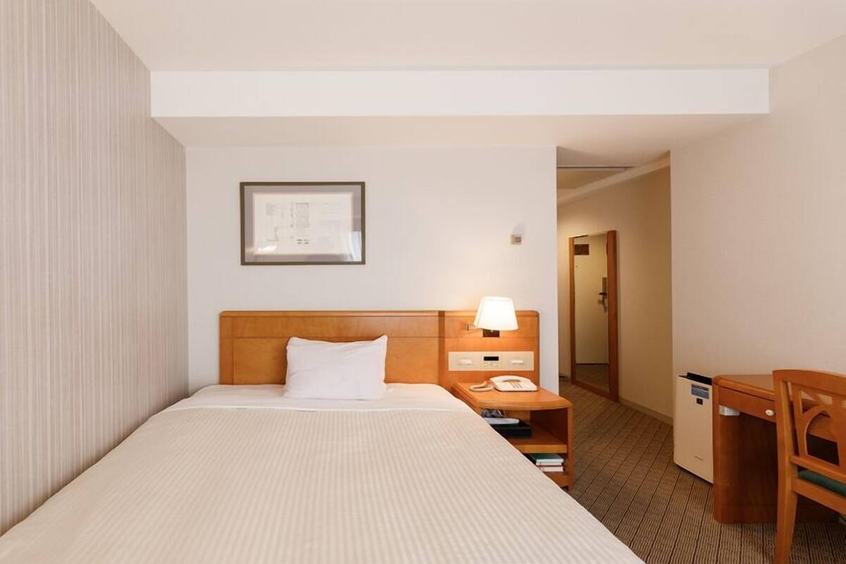 A hotel room with a single bed, a bedside table with a lamp, a phone, and a small shelf with books.