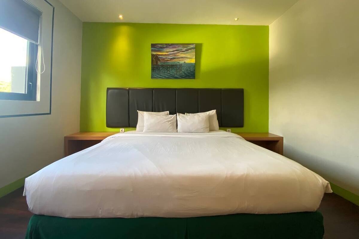 Habitación de hotel con una cama grande, paredes verdes, un cuadro sobre la cama y una ventana a la izquierda.