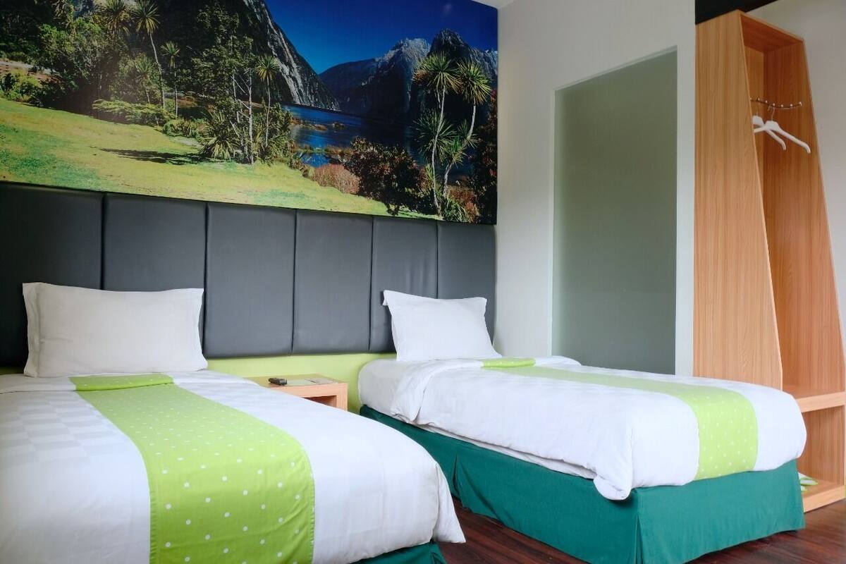 Habitación de hotel con dos camas, un cuadro grande de un paisaje, un armario de madera y una colcha verde.