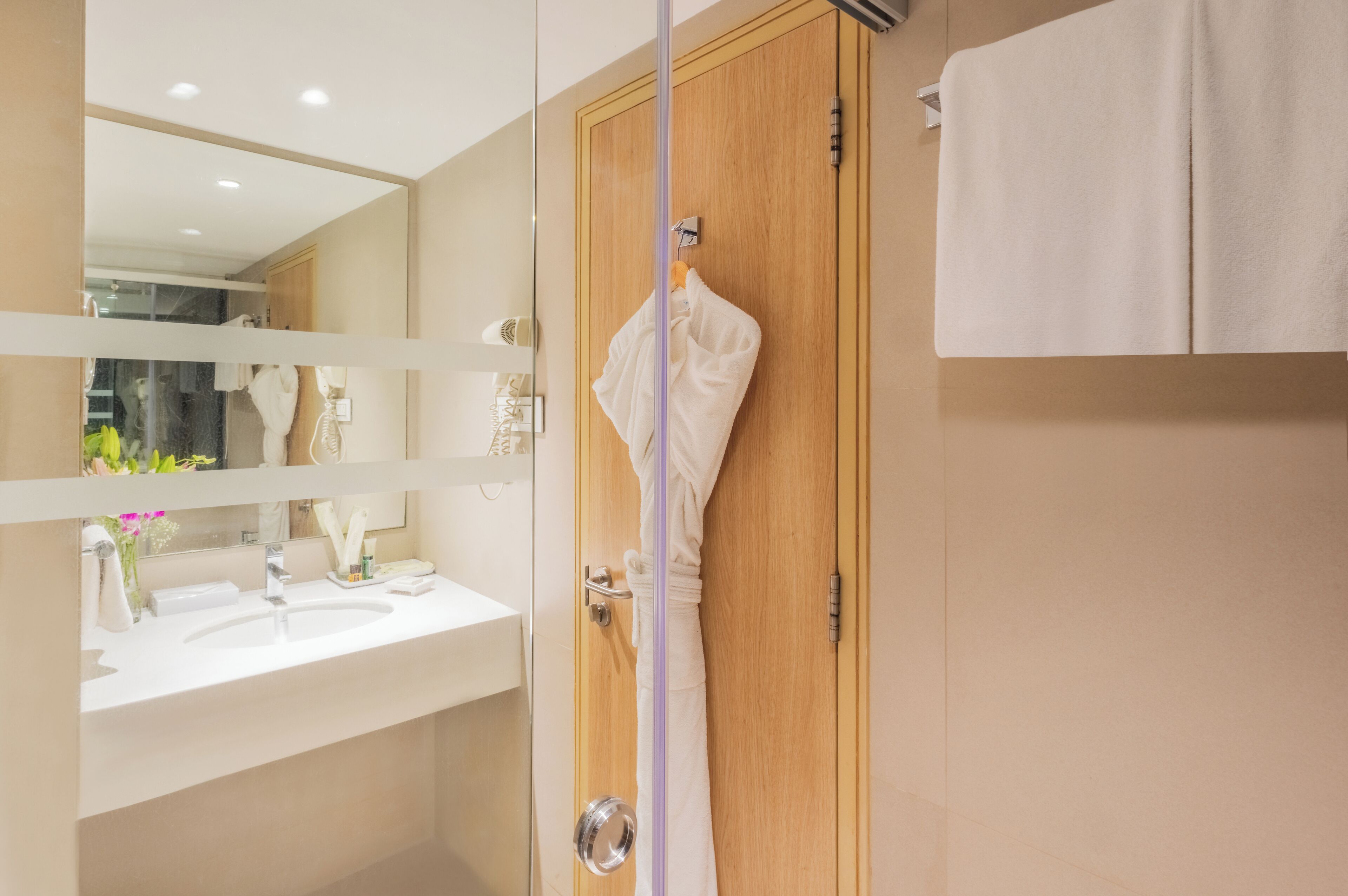 A bathroom with a wooden door, a white towel hanging on the door, a mirror, and a sink with a vase of flowers.