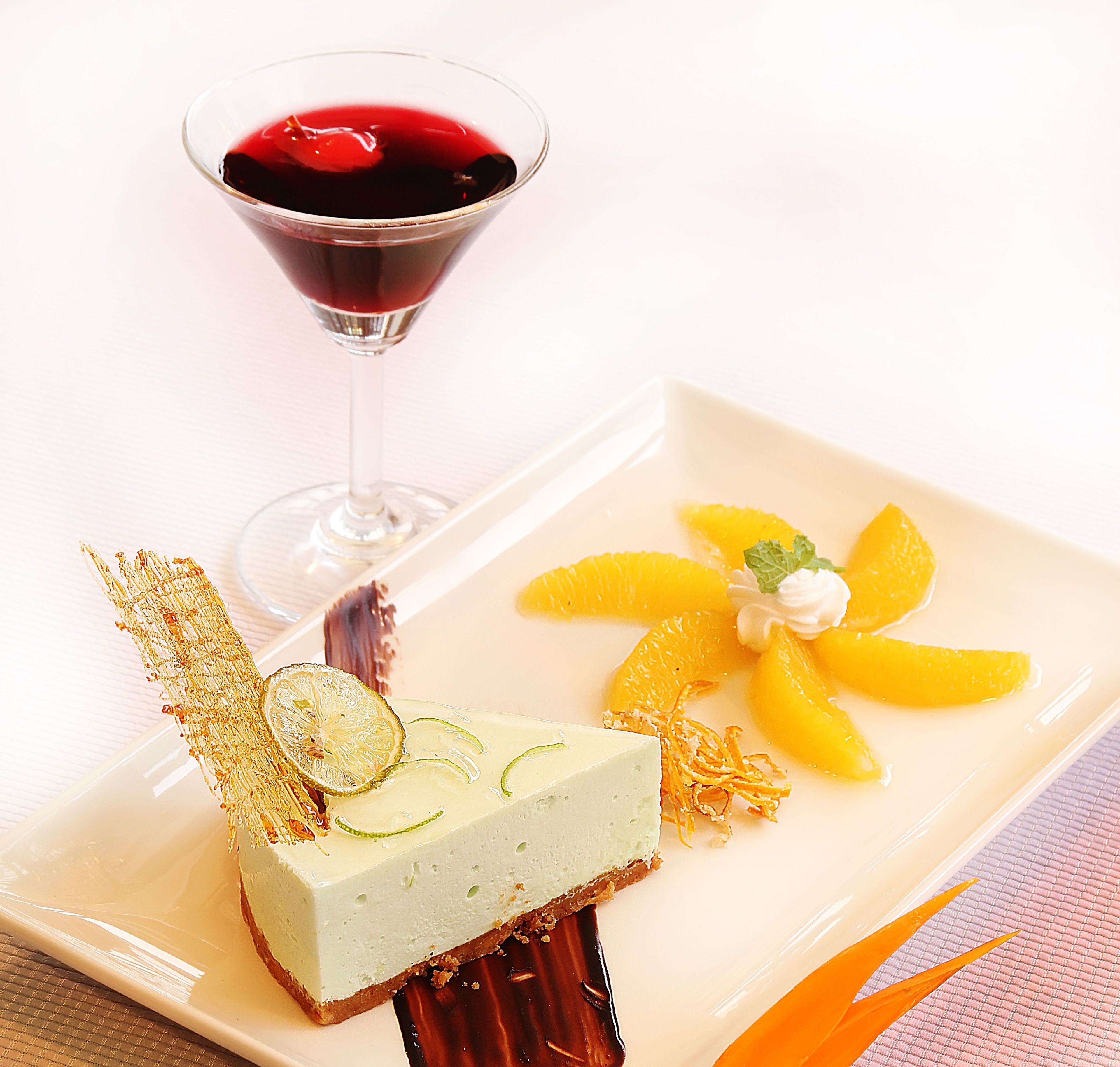 Une assiette à dessert avec une part de gâteau au fromage, des tranches d’orange et une boisson rouge servie dans un verre à martini.