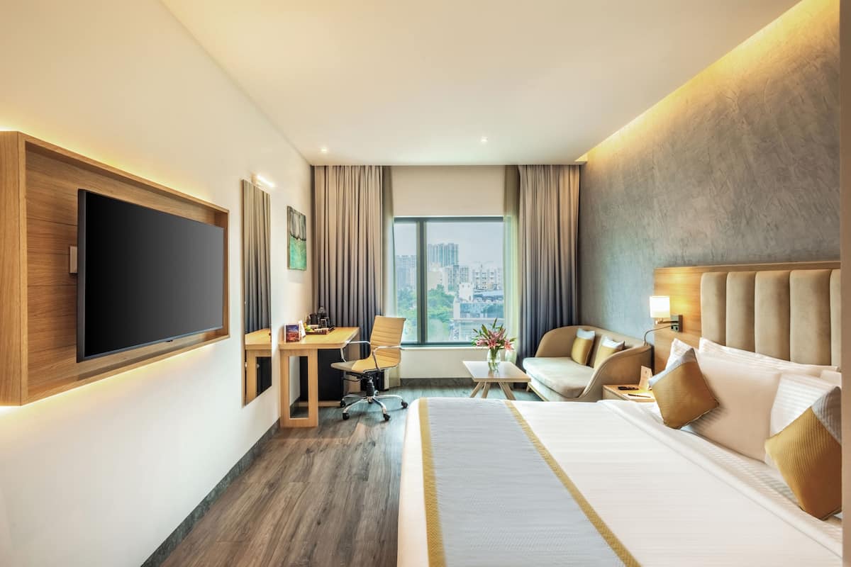 Habitación de hotel moderna con una cama grande, un escritorio, una silla, un televisor y vistas de la ciudad a través de amplios ventanales.