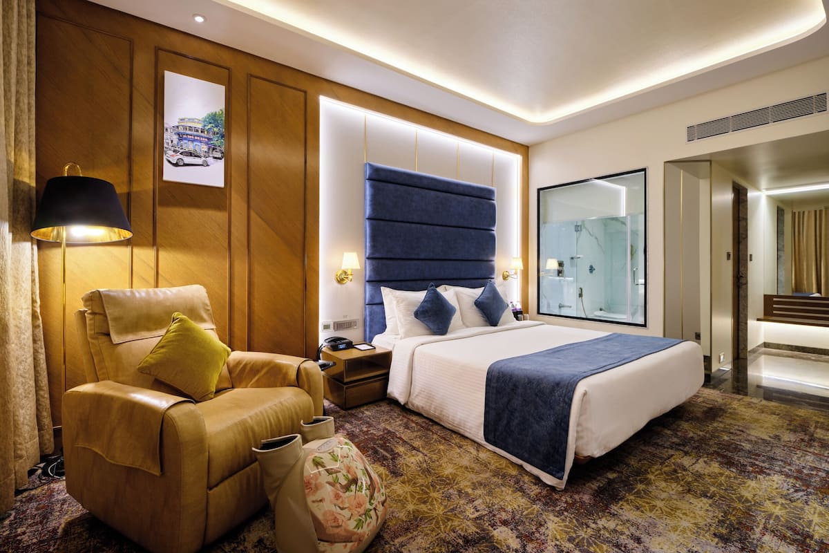 Una habitación de hotel moderna con una cama grande, un sofá, una mesita de noche y un baño visible a través de una puerta de cristal.