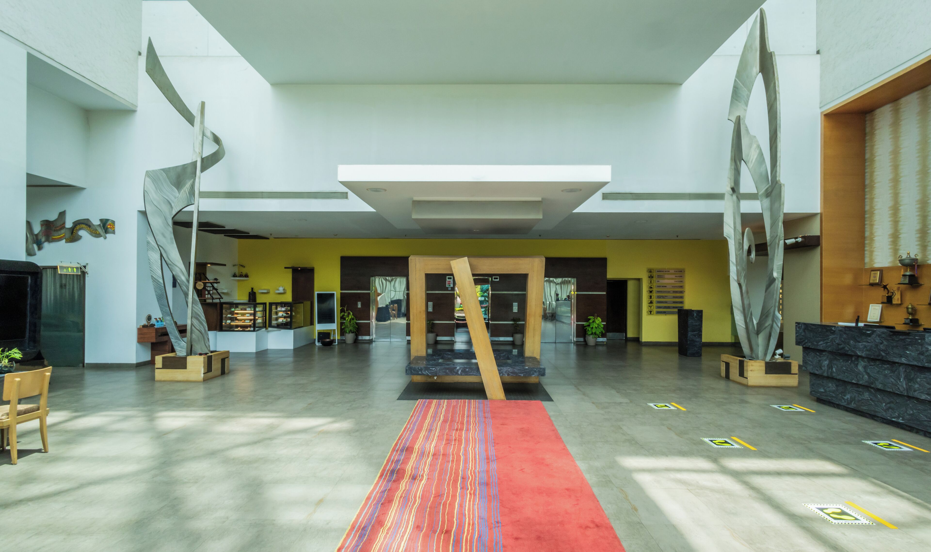 A spacious lobby with a central wooden sculpture, a reception desk, and a yellow wall with a signboard.