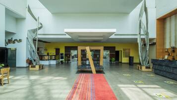A spacious lobby with a central wooden sculpture, a reception desk, and a yellow wall with a signboard.