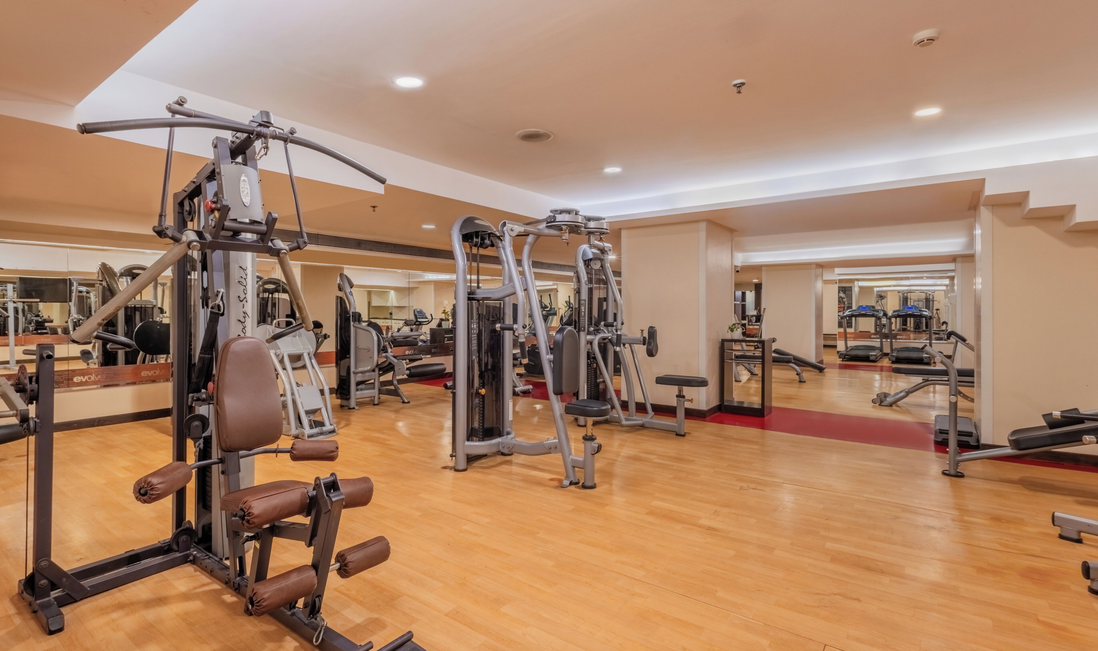 Un gimnasio amplio con diversas máquinas de ejercicio, suelo de madera y espejos en las paredes.