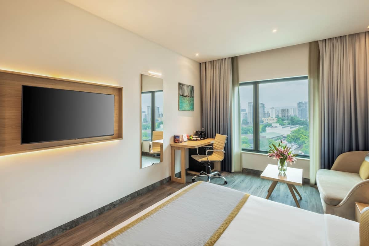 Habitación de hotel con una cama grande, un escritorio con silla, un televisor montado en la pared y vistas de la ciudad a través de amplios ventanales.