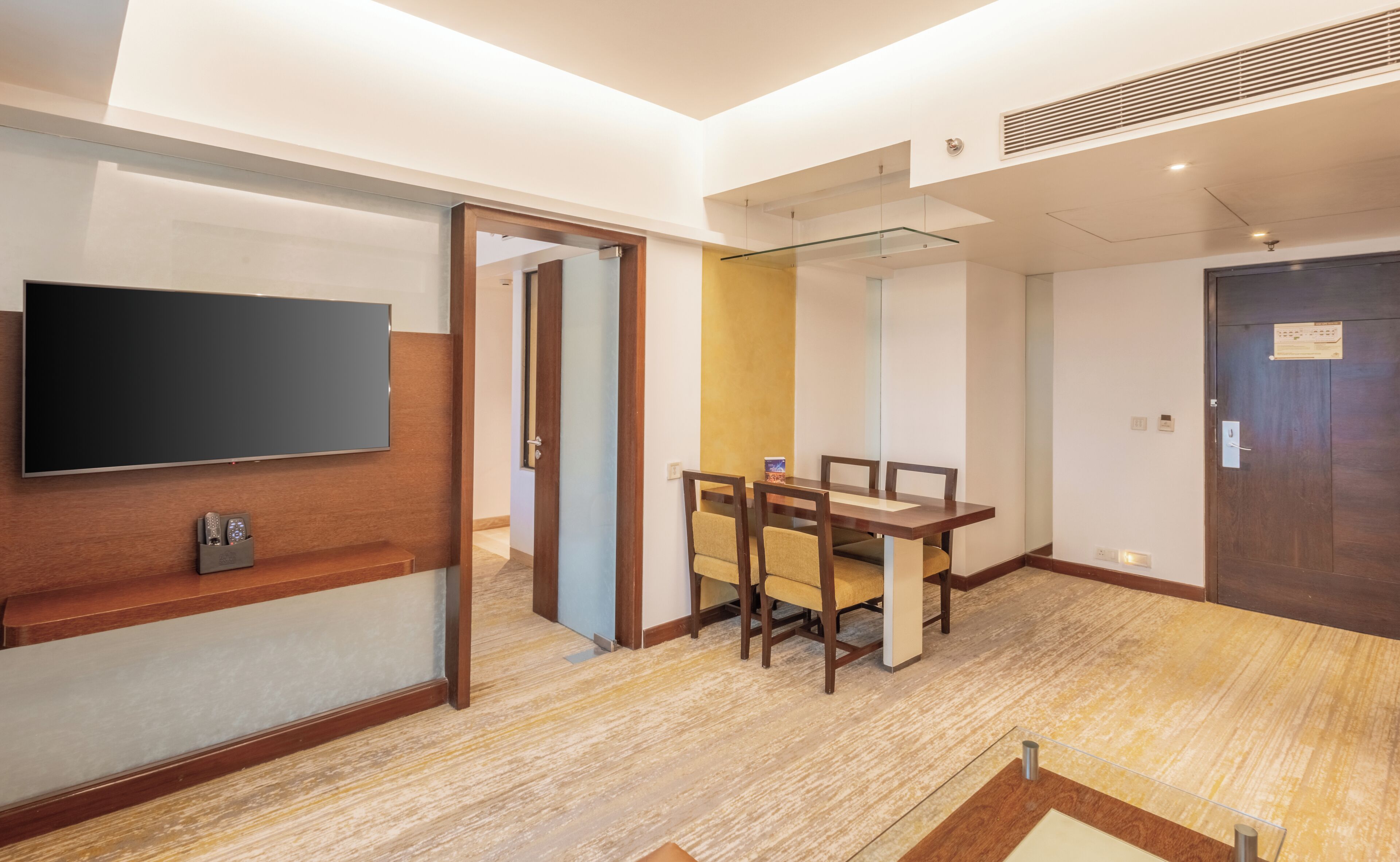 Quarto de hotel com televisão de tela plana na parede, escrivaninha de madeira com cadeira, e mesa de jantar com cadeiras.