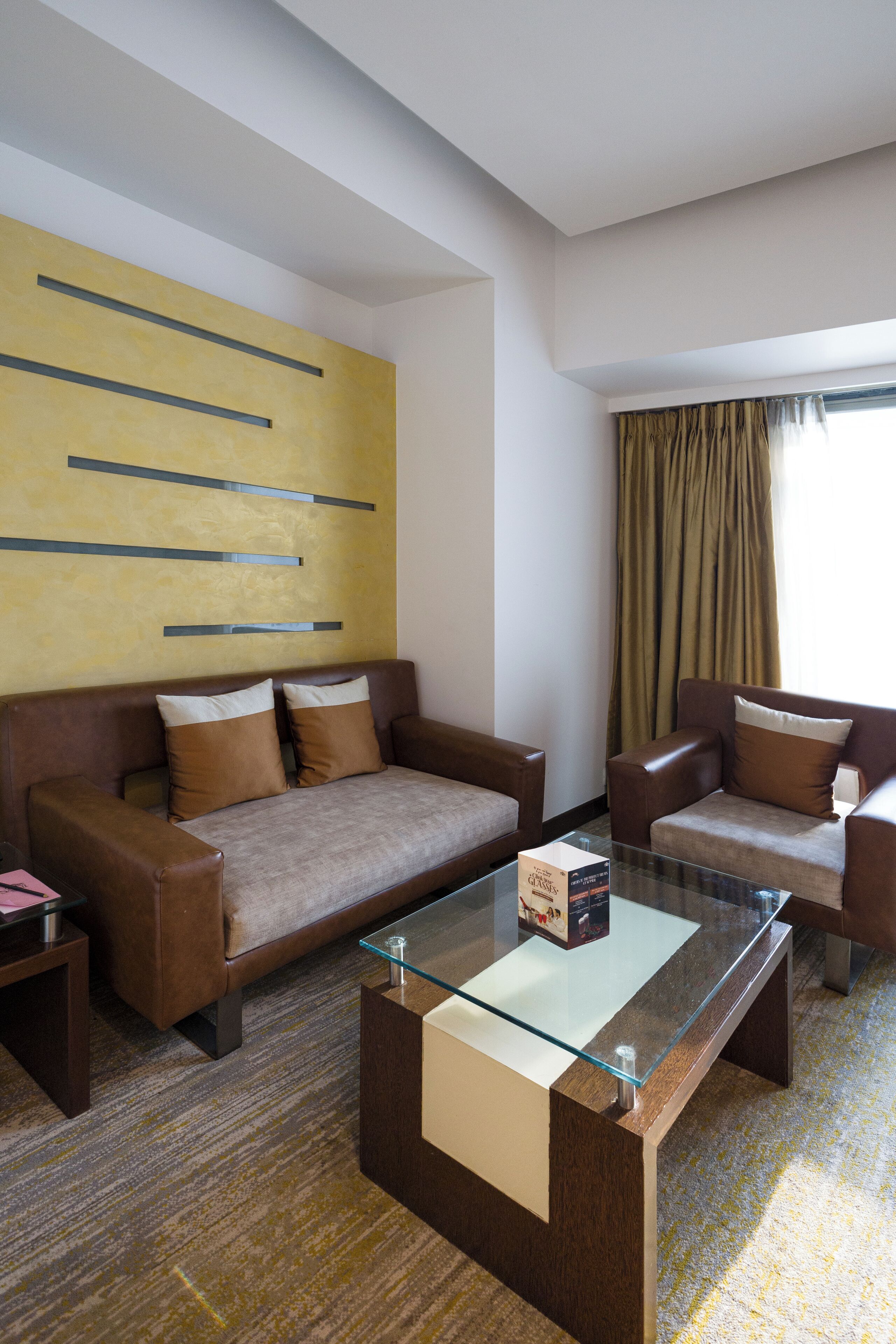Ein modernes Hotelzimmer mit einem braunen Ledersofa, einem Glastisch und einem großen abstrakten Gemälde an der Wand.
