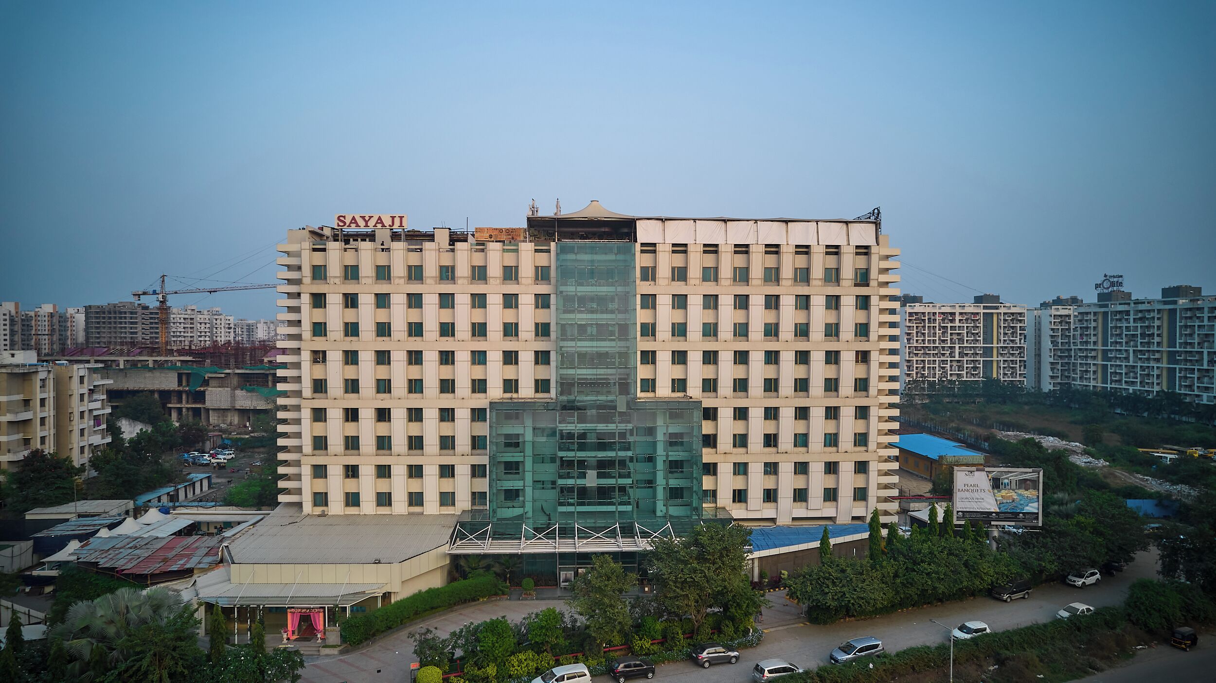A multi-story hotel with a central glass facade, a prominent 'SAYAJI' sign at the top, and a parking area in front.