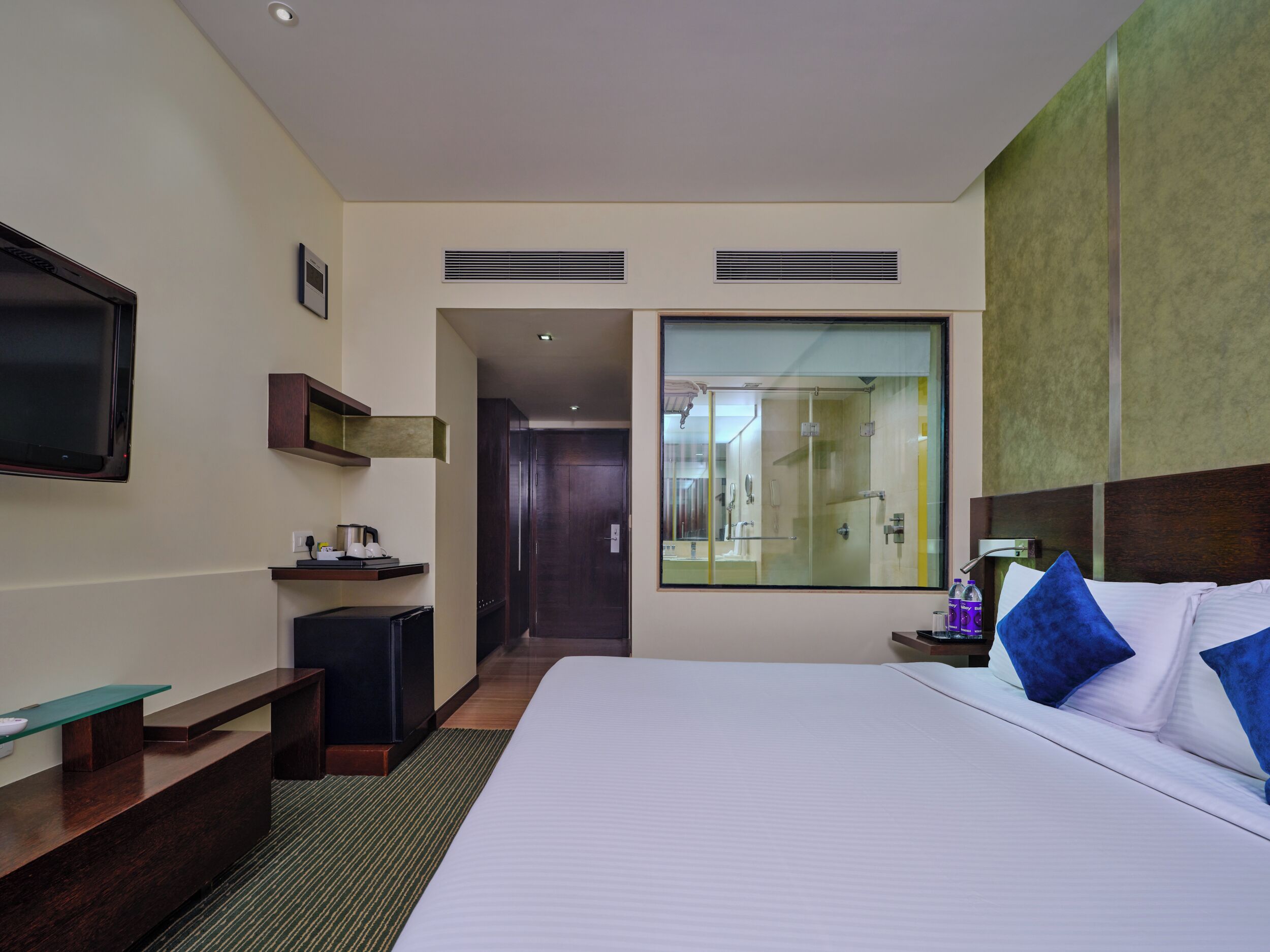 A hotel room with a large bed, a TV mounted on the wall, a small desk with a chair, a mirror, and a bathroom visible through the glass door.