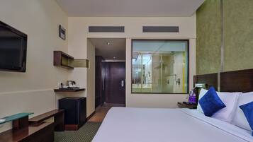 A hotel room with a large bed, a TV mounted on the wall, a small desk with a chair, a mirror, and a bathroom visible through the glass door.