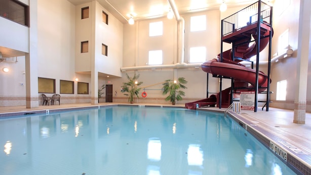 An indoor swimming pool with a waterslide, chairs, and a view of the sky through large windows.