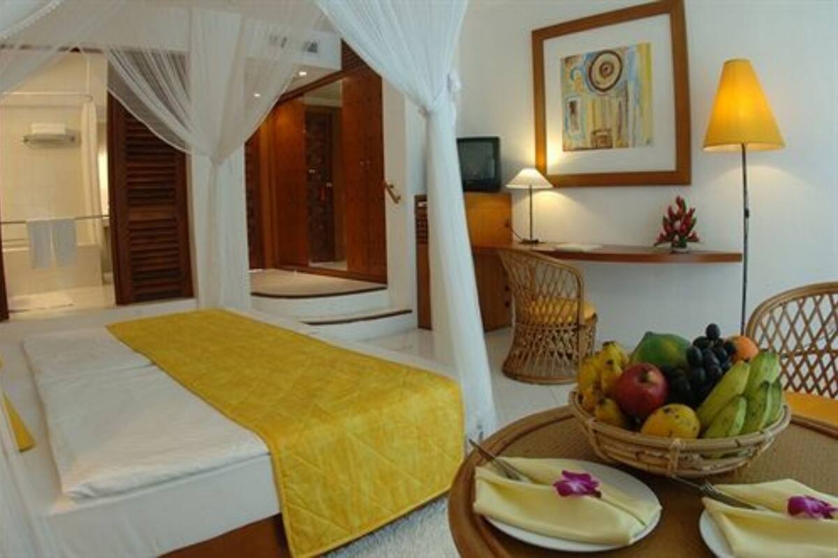 A hotel room with a bed, a bedside table with a fruit basket, a lamp, and a framed picture on the wall.