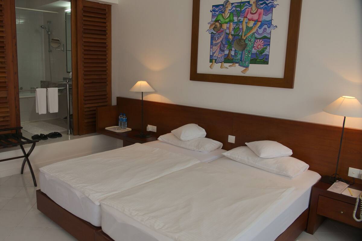 A hotel room with a double bed, two bedside tables, a nightstand, a mirror, a shower area, and a towel rack.