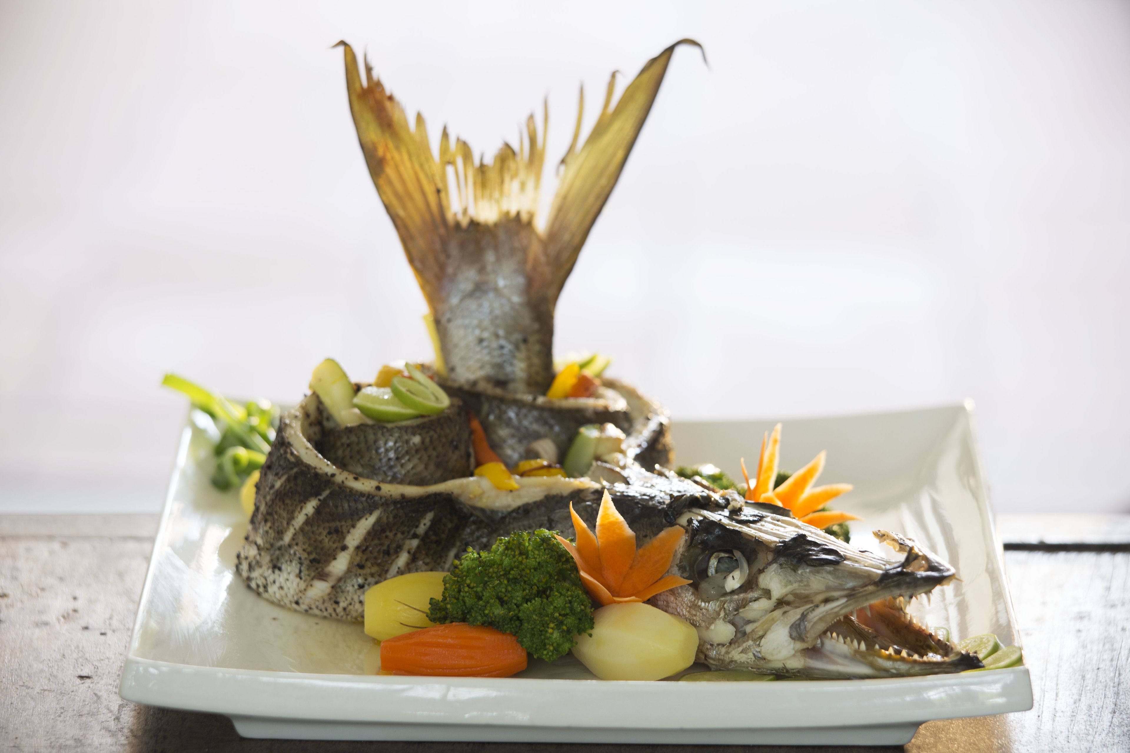 A white plate with a whole fish, vegetables, and a decorative leaf.