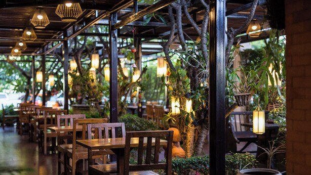 Restaurante interno com mesas e cadeiras de madeira, lanternas penduradas e plantas em vasos.
