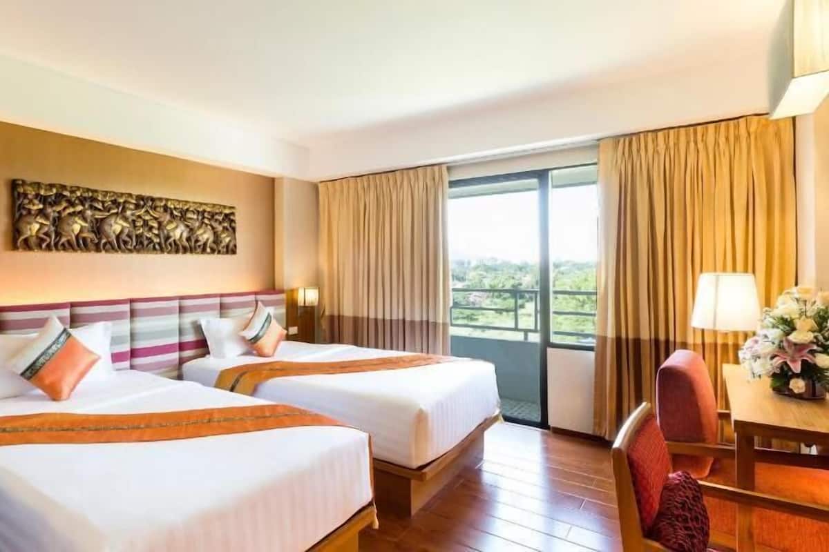 A hotel room with two beds, a dining table, a chair, a lamp, and a view of greenery.