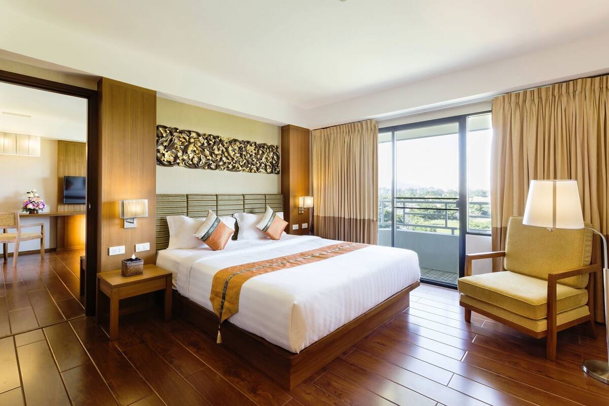 A hotel room with a large bed, a sitting area with a chair, a television, and a balcony with curtains.