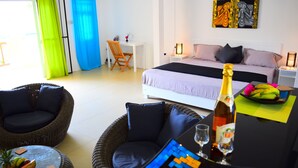 Ett hotellrum med en säng, ett skrivbord, en fåtölj i rotting, ett litet bord med en flaska och glas, och ett färgstarkt konstverk på väggen.
