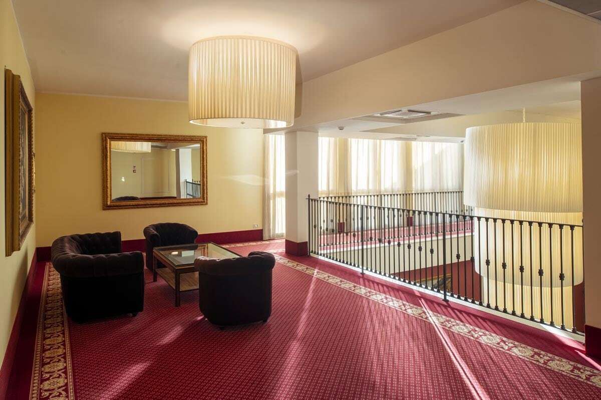 A hotel room with a red carpet, a sofa, a chair, a table, a mirror, and a balcony with a railing.