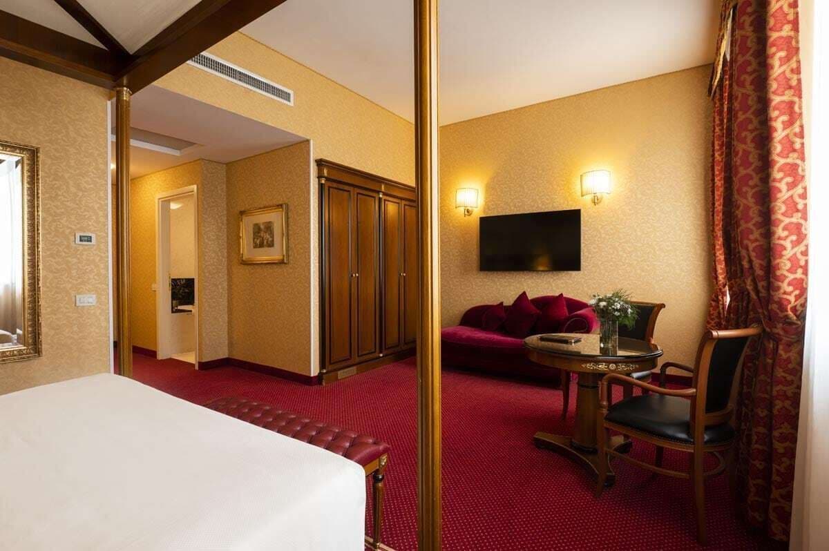 Una habitación de hotel con una cama, alfombra roja, televisión, sofá y una mesita con un jarrón.