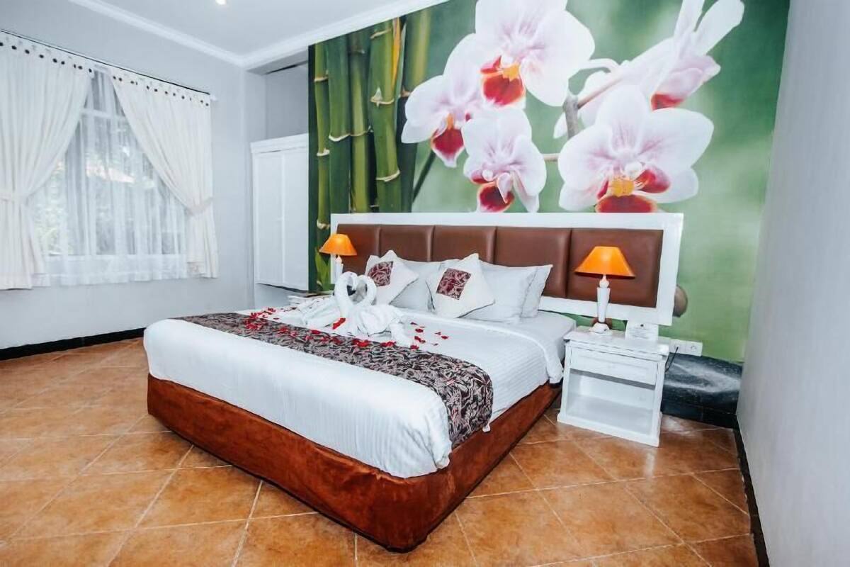A bedroom with a bed, two bedside lamps, a nightstand, a wall with an orchid mural, and a window with white curtains.