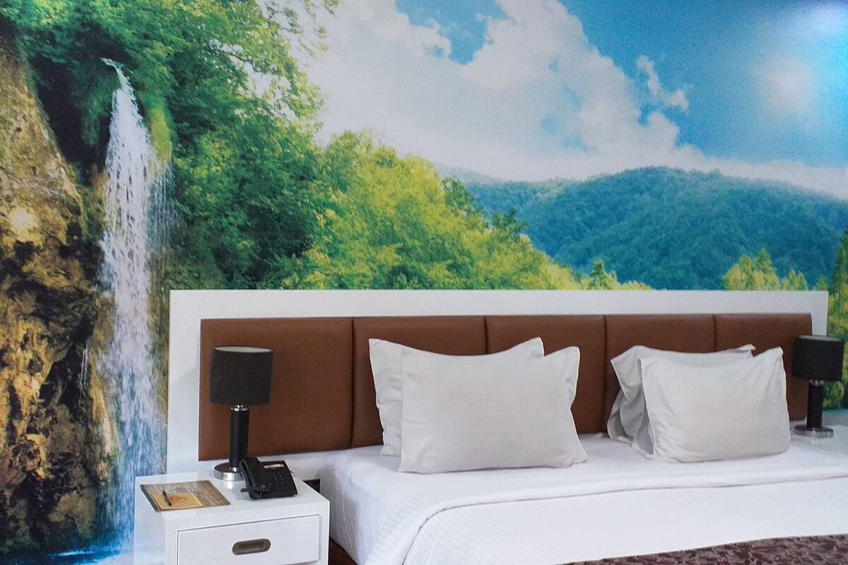 A hotel room with a bed, white bedding, a nightstand with a lamp, a phone, and a painting of a waterfall and forest.