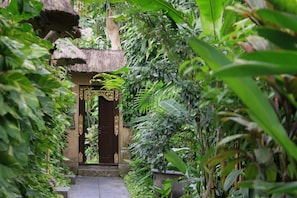 A narrow pathway leading to a traditional building with a thatched roof and ornate door, surrounded by lush greenery.