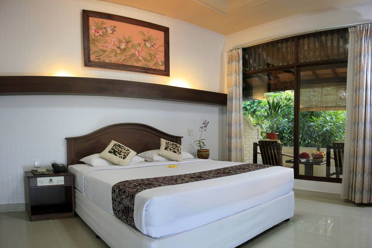 A hotel room with a bed, a nightstand, a potted plant, and a view of the outdoors.