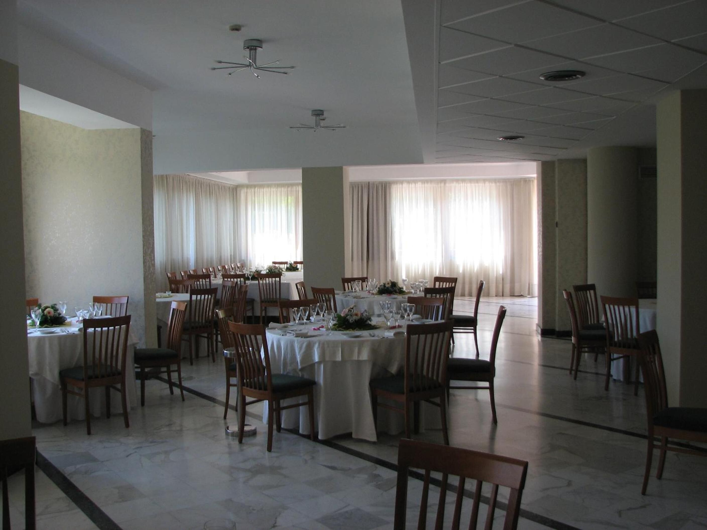 Une salle à manger spacieuse, dotée de nombreuses tables et chaises, de grandes fenêtres avec des rideaux et d’un ventilateur de plafond.