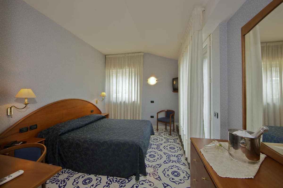 Una habitación de hotel con cama, mesita de noche, lámpara, silla y un espejo grande.