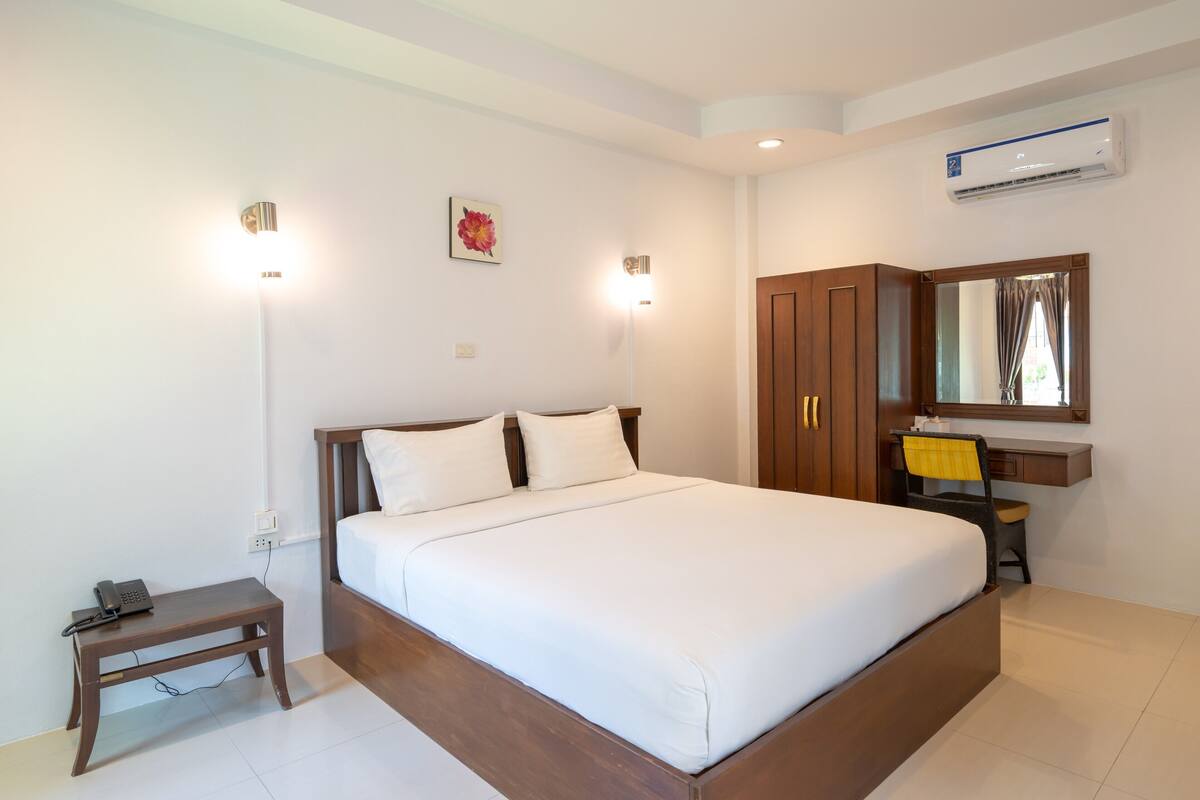 A bedroom with a large bed, bedside lamps, a wooden nightstand, a desk with a chair, and an air conditioning unit.