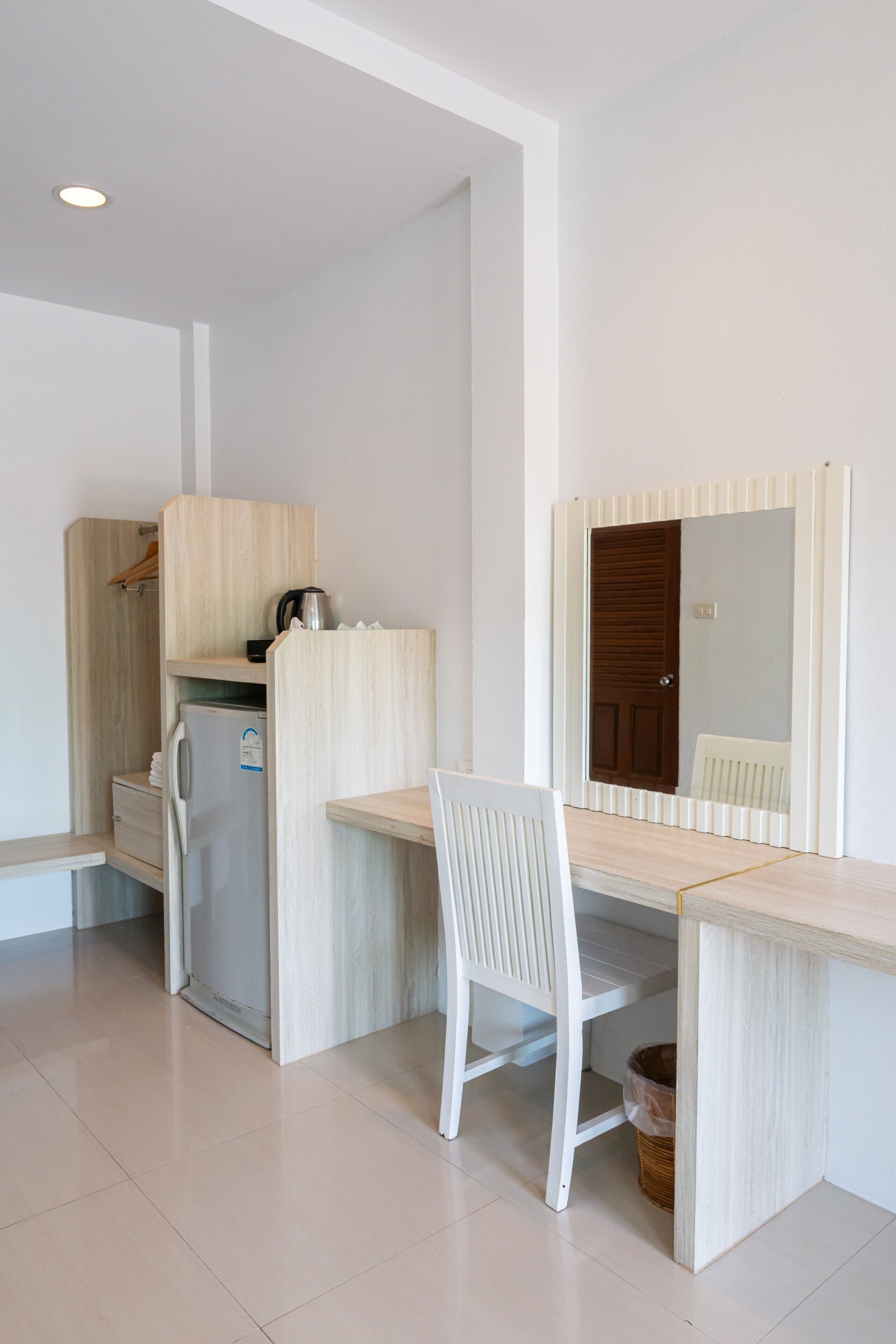 A compact room with a kitchenette, a white chair, a wooden desk, and a mirror.