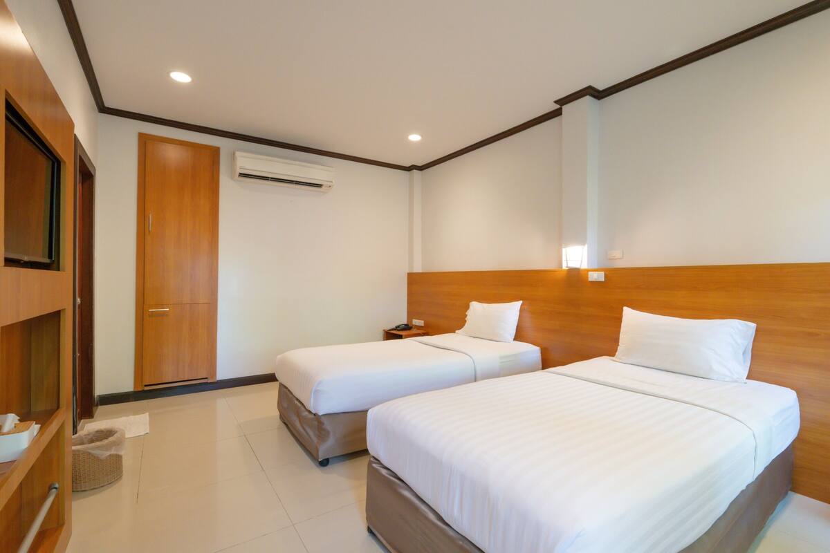 A hotel room with two beds, a wooden headboard, an air conditioning unit, a bedside table, and a wooden wardrobe.