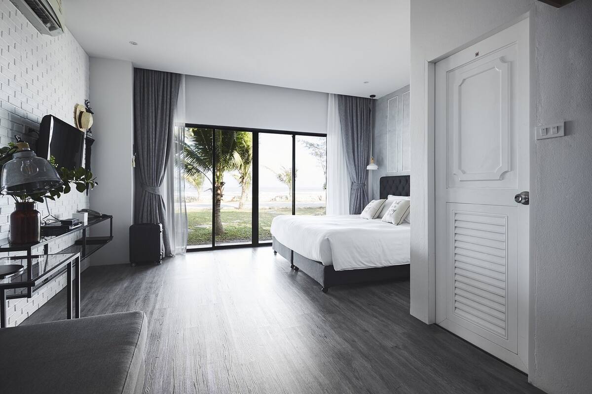 A modern hotel room with a large bed, a desk with a lamp, a sliding glass door to the outside, and a view of palm trees.