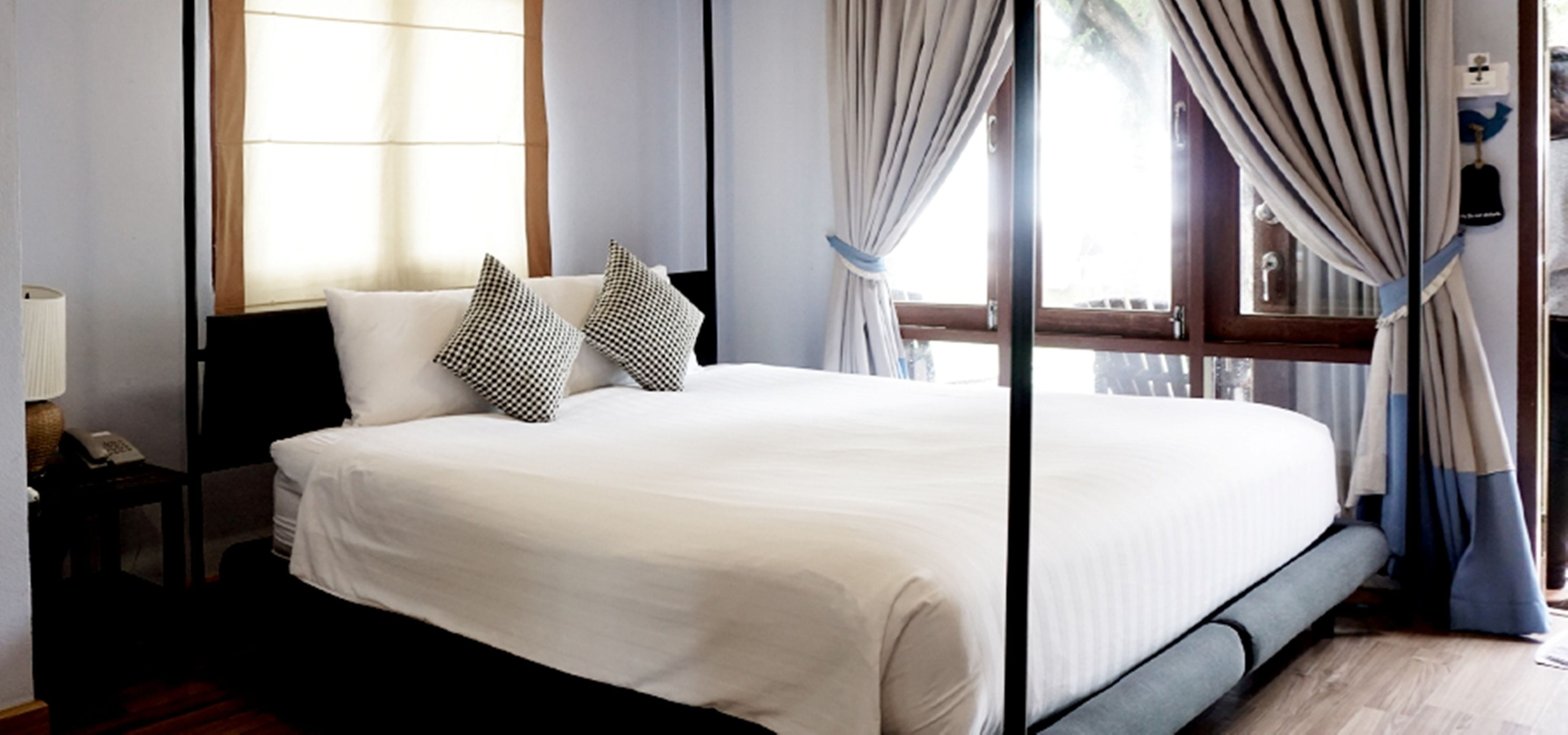 A neatly made bed with white bedding and two patterned pillows, a bedside table with a lamp, and a window with curtains.