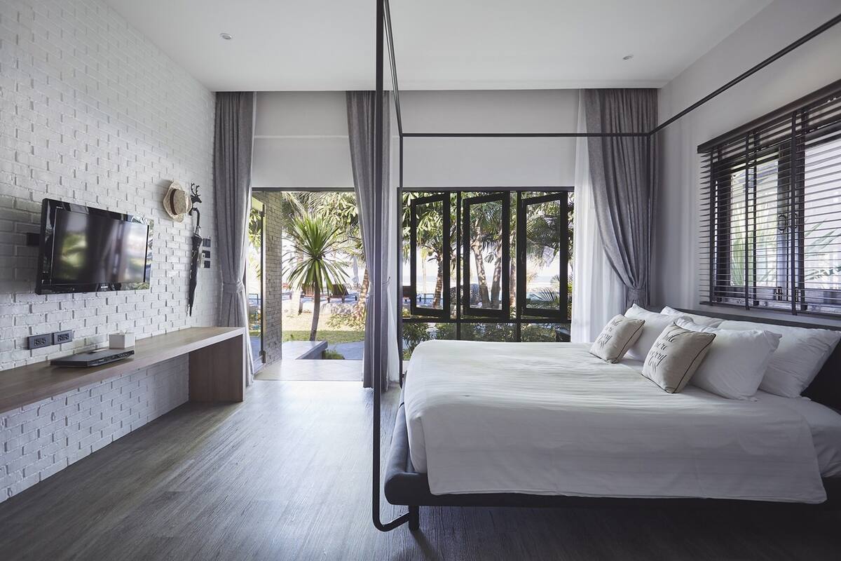 A modern bedroom with a large bed, a TV mounted on the wall, and a view of palm trees through the window.