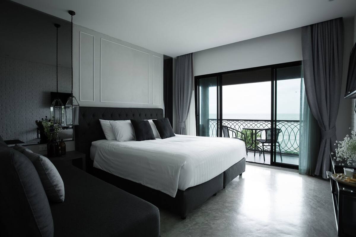 A modern bedroom with a large bed, a sofa, a TV, and a balcony with a view.