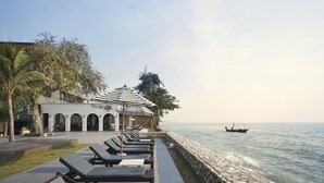 A seaside resort with lounge chairs along a stone wall, a small boat on the water, and a building with arched windows. - SiamBeach Resort (Cha-am)