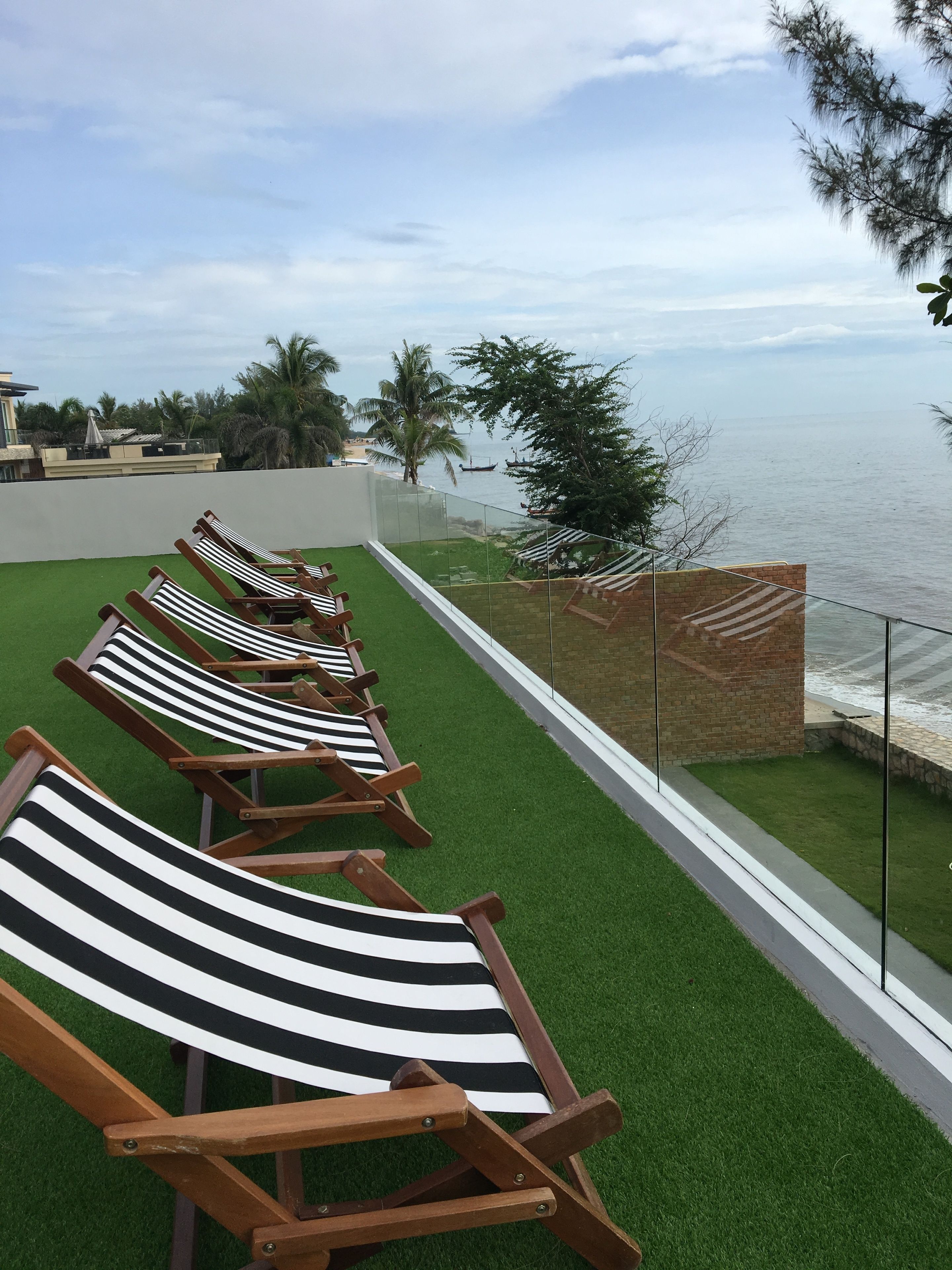 A row of striped lounge chairs on artificial grass, overlooking a body of water with boats and palm trees in the background.