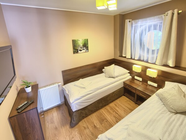 Une chambre d’hôtel avec deux lits simples, une petite table de chevet avec une lampe, une plante en pot, un radiateur et une fenêtre avec des rideaux.